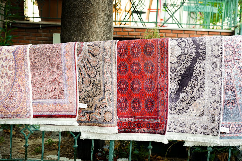 Multiple Persian-style rugs are draped over a metal railing outdoors, displaying intricate patterns and vibrant colors including reds, beiges, black, and blue. The rugs are clean and well-maintained, with visible fringes along the edges. Behind the rugs, there is a large tree trunk providing shade, and a background of a brick wall, green plants, and a metal fence. The scene is illuminated by natural daylight, highlighting the detailed designs and the pristine condition of the rugs. This setting suggests professional domestic cleaning or surface cleaning services offered by Stockwell Carpet Cleaning, specializing in deep cleaning and sanitisation of rugs and decorative floor coverings in residential environments.
