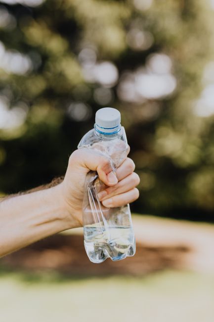 A close-up of a person's hand gripping a clear plastic water bottle filled with water, held outdoors against a blurred background of green trees and sunlight. The scene focuses on the hand and bottle, with natural light highlighting the transparent surface and the blue cap. The background suggests a park setting, aligning with the theme of cleaning and outdoor maintenance, as emphasized by the website stockwellcarpetcleaning.com and the service page titled 'Brockwell Park rug cleaning with eco disposal, STOCKWELL.' The clean, fresh appearance of the water and the outdoor environment reflect hygiene and eco-friendly practices associated with domestic and commercial cleaning services offered by Stockwell Carpet Cleaning.