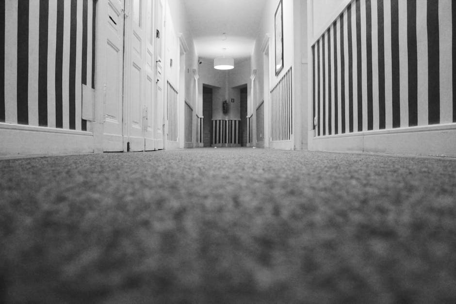 A close-up, black-and-white photograph of a hallway with a textured carpet floor. The walls are decorated with vertical striped wallpaper in alternating light and dark shades. Multiple white doors with panel designs line both sides of the corridor, leading towards a brightly lit area with a ceiling light fixture visible at the far end. This setting showcases a clean, well-maintained space typical of residential or commercial interiors, emphasizing surface cleanliness and detailing, consistent with professional deep cleaning and sanitisation services possibly offered by Stockwell Carpet Cleaning.