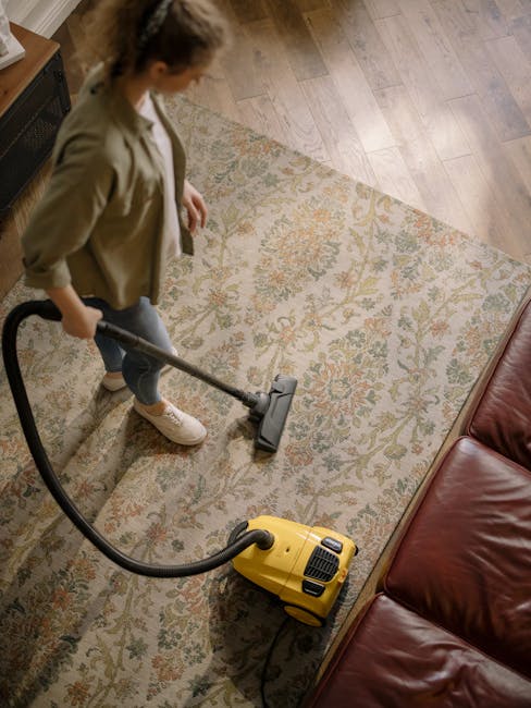 A person using a yellow vacuum cleaner to deep clean a patterned area rug in a living room with wooden flooring. The individual is wearing casual clothing, including a beige jacket and white sneakers, and is focused on removing dirt from the soft, intricate design of the rug. The room features a red leather sofa nearby, and the scene is well-lit, highlighting the cleanliness of the carpet and the surrounding space. This image showcases surface cleaning and deep cleaning techniques offered by Stockwell Carpet Cleaning for domestic settings.