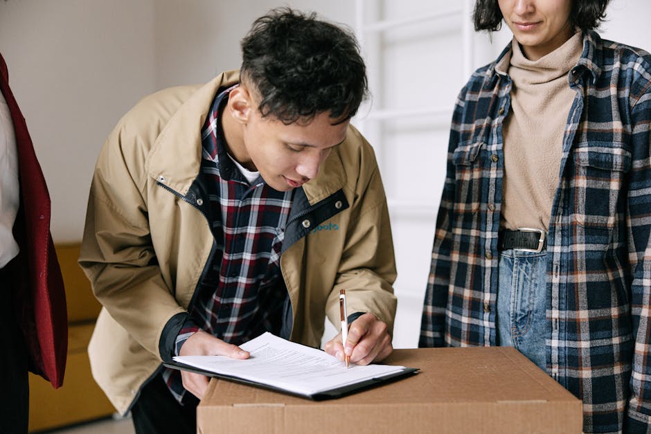 A person in a beige jacket and checked shirt is signing a document on top of a brown cardboard box, with another person standing beside them wearing a blue plaid shirt and beige turtleneck. The setting appears to be an indoor residential space with neutral-colored walls and natural light. The scene suggests a formal handover or inspection related to end of tenancy cleaning, emphasizing surface cleaning, hygiene, and maintenance in a domestic environment, aligning with the services offered by Stockwell Carpet Cleaning. Visible surfaces include the cardboard box, the person's hands, and part of the wall and furniture in the background, highlighting a clean and organized space.