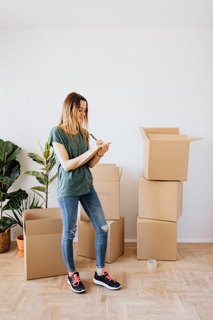 A young woman with shoulder-length blonde hair, dressed in a casual green T-shirt and ripped blue jeans, standing inside a room with a light-colored wooden floor and plain white wall. She is holding a clipboard and pen, appearing to be inspecting or taking notes on the cleaning process. Around her, there are several cardboard moving boxes, some stacked and one open at the top, indicating a relocation or deep cleaning scenario. A large potted plant with broad green leaves is positioned behind her on the left side, adding a touch of greenery to the space. The room is well-lit with natural light, emphasizing the cleanliness and readiness for surface cleaning or sanitisation, consistent with a professional end of tenancy cleaning service. This image is associated with Stockwell Carpet Cleaning on their website's end of tenancy cleaning checklist for Stockwell Park Estate.