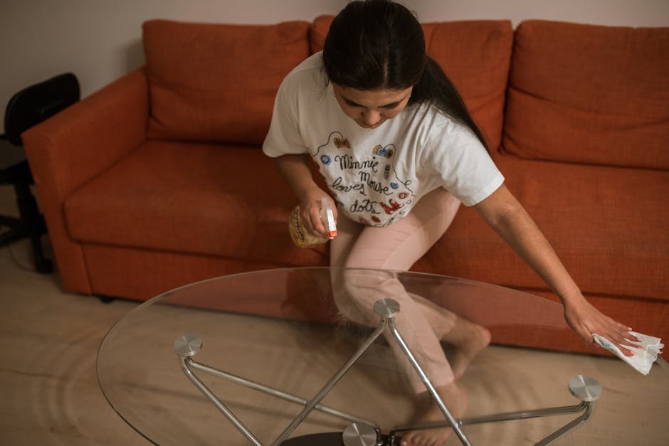 A woman with dark hair, wearing a white T-shirt and light pink pants, is engaged in domestic cleaning on a glass coffee table in a living room. She is kneeling with one knee on the floor, using a cloth in her right hand to wipe the surface of the transparent, round glass table, which has a metal frame with four supports. The room features a plush, orange sofa with matching cushions positioned against a wall. The lighting appears soft and warm, emphasizing the cleanliness and shine of the glass surface, which is free of dust or smudges. A black chair is partially visible behind the sofa, and the overall setting indicates a tidy, well-maintained space where surface cleaning and sanitisation are carried out to ensure hygiene at home. The scene highlights domestic deep cleaning efforts by Stockwell Carpet Cleaning, as part of their same-day sofa cleaning services in a busy household environment.
