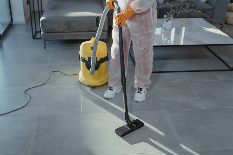A person wearing white protective coveralls and yellow rubber gloves is using a vacuum and a cleaning wand to perform surface cleaning on large, gray ceramic floor tiles in a modern living room. The room features a gray sofa with decorative pillows, a glass-top coffee table, and a black metal side table, all arranged on the clean, shiny tiled floor. The individual operates a yellow commercial vacuum cleaner with a hose attachment, and the scene is well-lit with natural light, highlighting the cleanliness and maintenance process. Stockwell Carpet Cleaning specializes in deep cleaning and sanitisation for residential spaces, ensuring hygienic and spotless surfaces in busy homes.